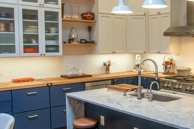 Kitchen by Modern Kitchen with navy base cabinets, a butcher block counter, white glass-front uppers and a marble-look island with a farmhouse sink