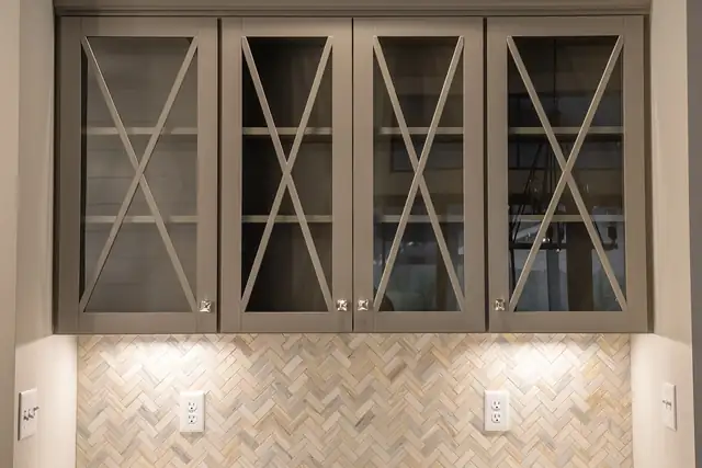 Taupe wall cabinets with X-pattern glass doors over a herringbone stone backsplash